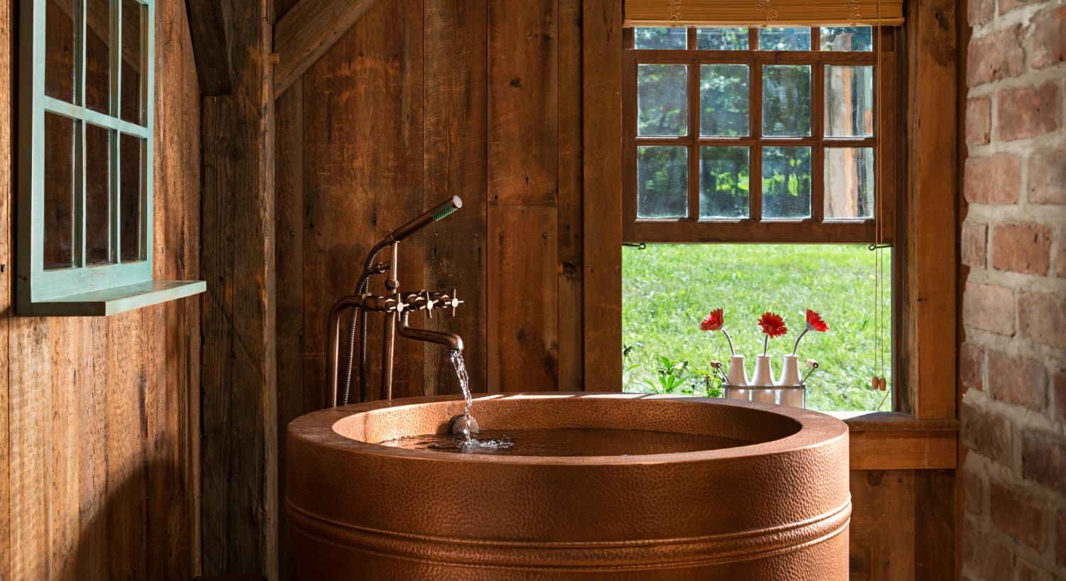 Wood and brick room with round copper tub, antique faucet and open window with three white vases of red flowers