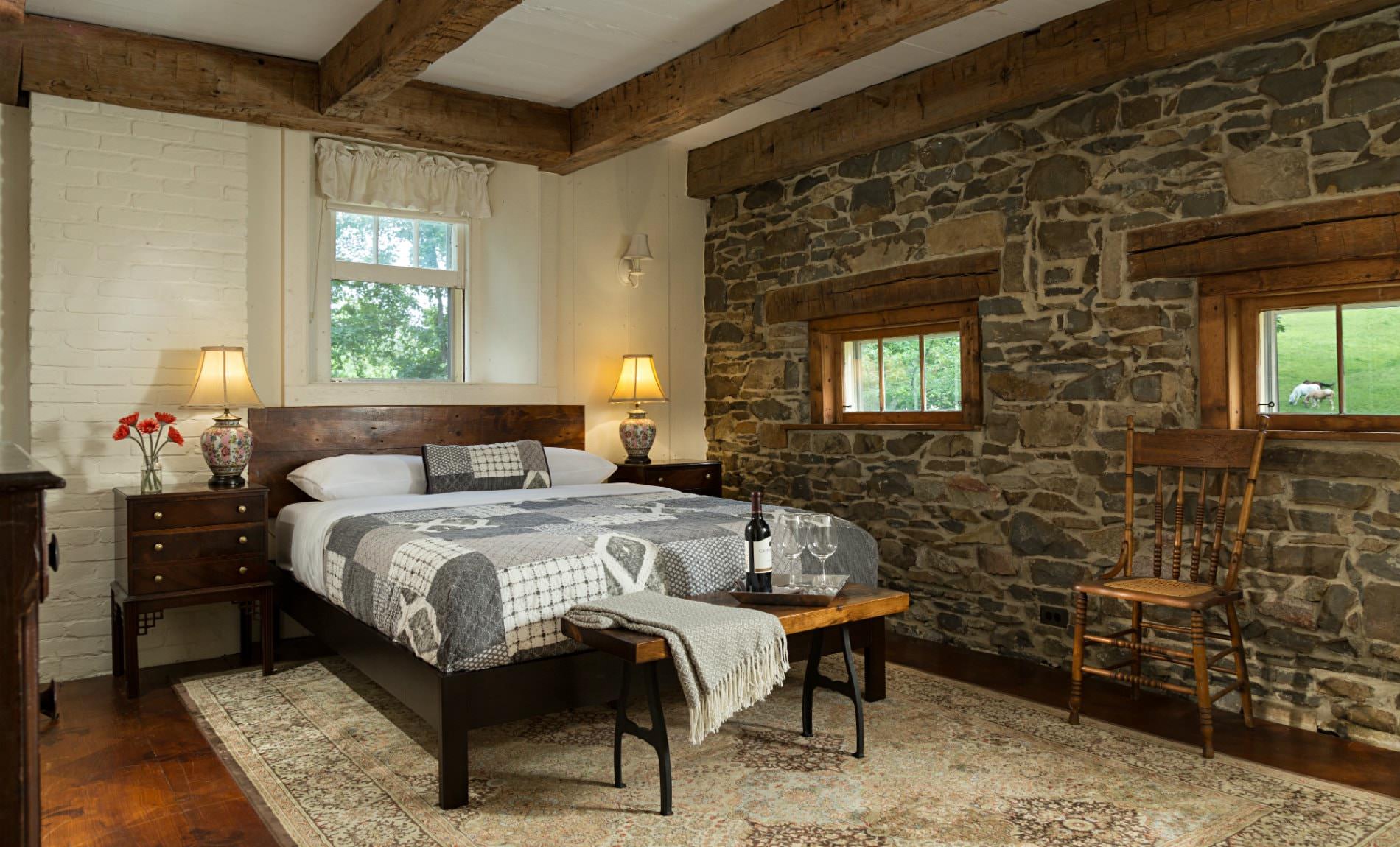 Spacious white room, exposed rustic beams, stone wall with two small windows, light colored rug, gray and white covered bed