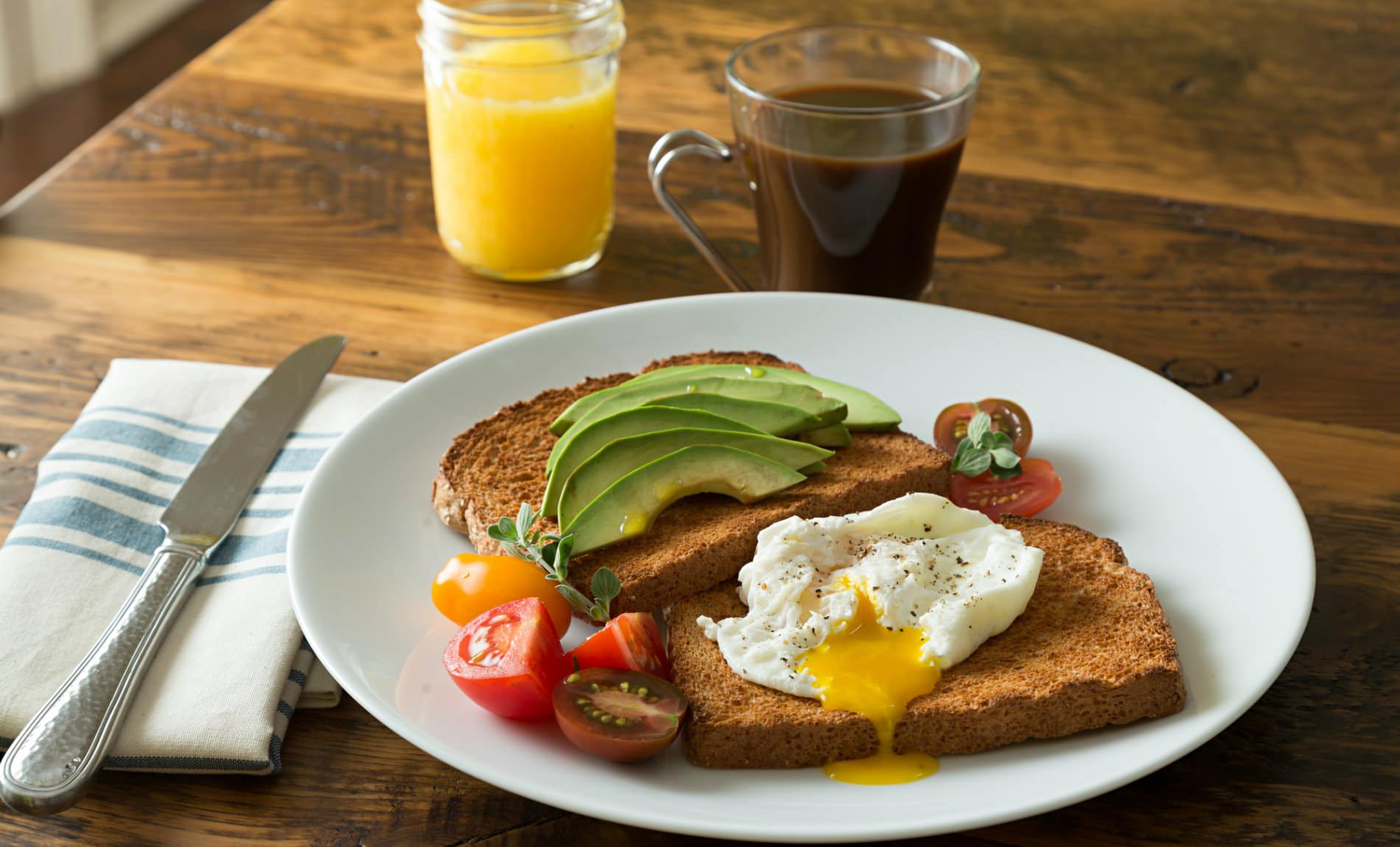 White plate of toast, avocado slices, fried egg and sliced cherry tomatoes, glass of juice and cup of coffee