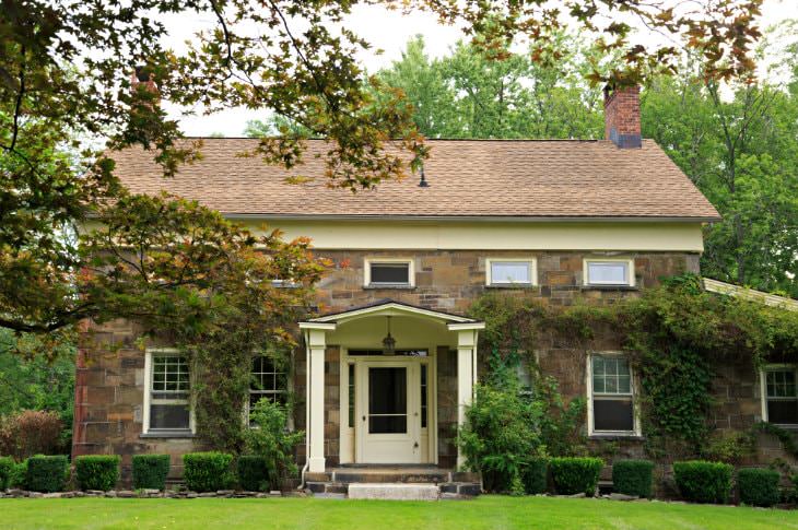 Front exterior view of brown stone inn with covered gable entry, brown shingled roof surround by lawn and trees