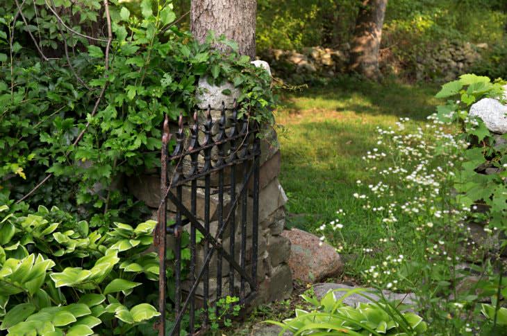 Stone base with a black wrought iron gate surrounded by grass, plants, bushes and trees