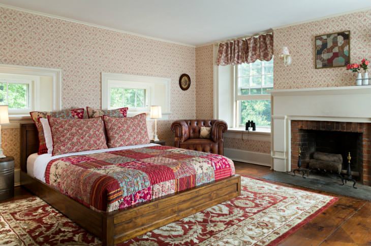 Spacious cozy room with warm wood floors, several windows, red floral quilted bed, fireplace and brown leather club chair