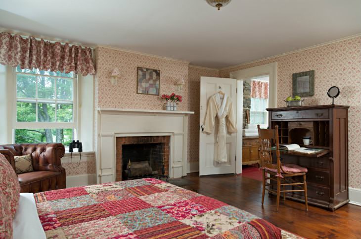 Spacious cozy room with warm wood floors, large windows, red floral quilted bed, fireplace and antique secretary