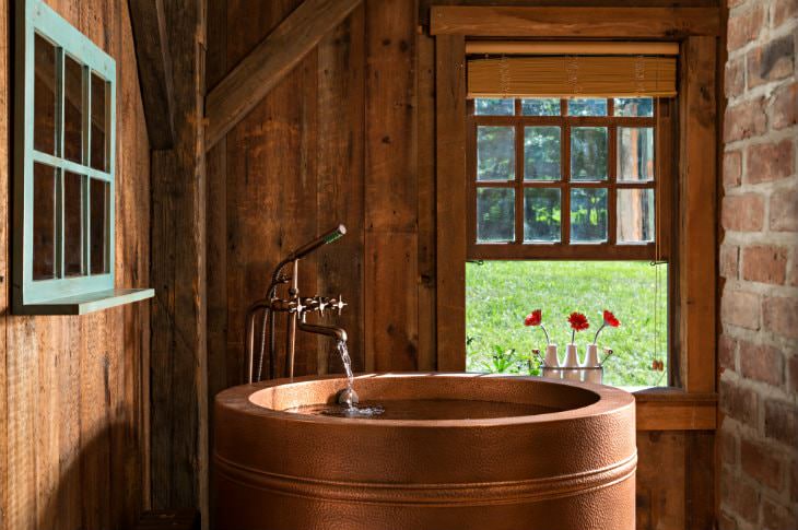 Wood and brick room with round copper tub, antique faucet and open window with three white vases of red flowers