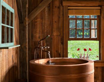 Wood and brick room with round copper tub, antique faucet and open window with three white vases of red flowers