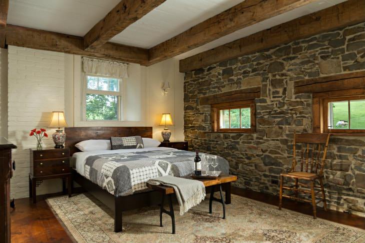 Spacious white room, exposed rustic beams, stone wall with two small windows, light colored rug, gray and white covered bed