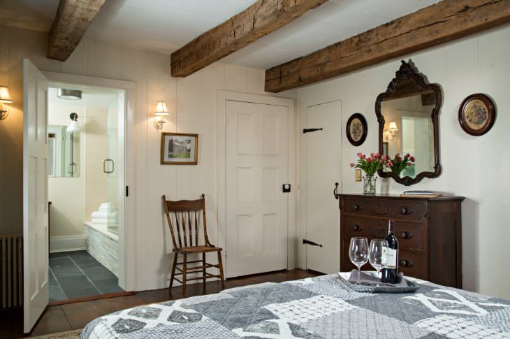 White room, exposed rustic beams, gray and white covered bed topped with wine bottle and wine glasses on a tray