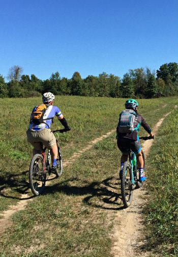 Man and woman riding bikes along dirt path in an open meadow surrounded by trees and blue skies