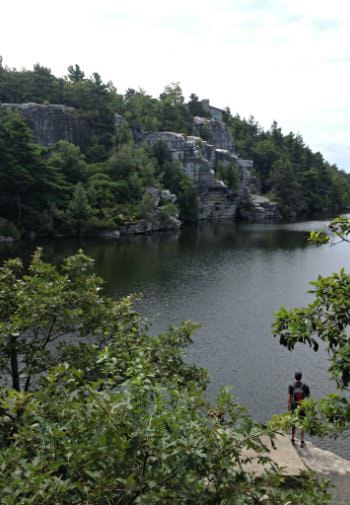 Man standing on stone ledge overlooking body of water surrounded by hills covered in rock and trees