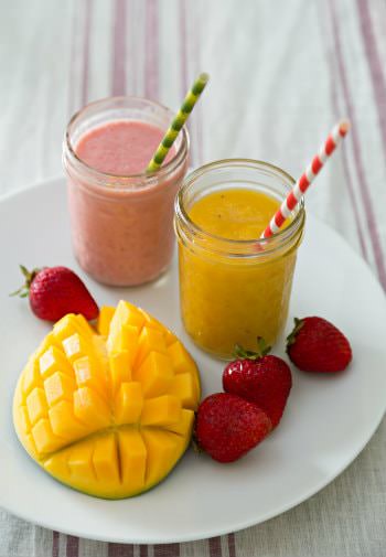 White plate topped with fresh mango and strawberries and two mason jar smoothies with green and red striped straws