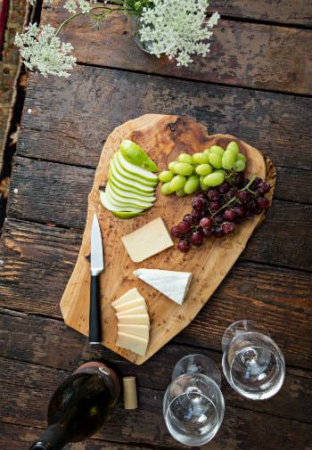 Rustic table topped with cheese board with grapes and apple slices, bottle of uncorked wine and two wine glasses