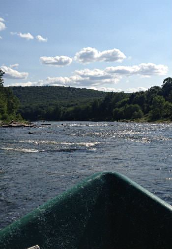Tip of green canoe in body of water surrounded by green trees and hills, blue skies and white clouds