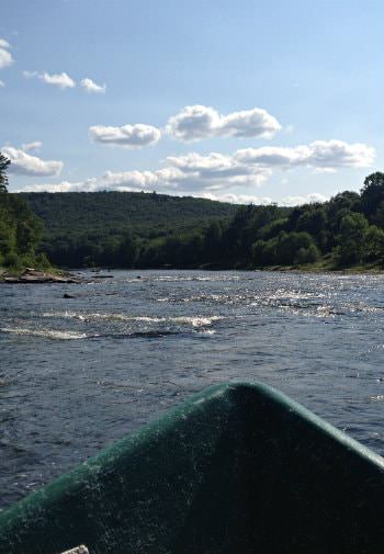 Tip of green canoe in body of water surrounded by green trees and hills, blue skies and white clouds