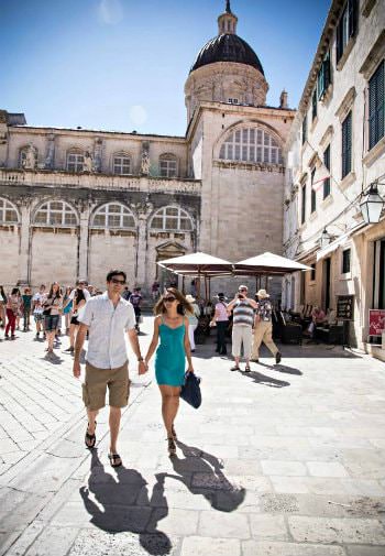 Man and woman holding hands walking through stone courtyard amidst a crowd of people and old stone buildings