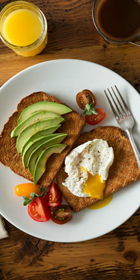 White plate topped with toast, avocado ,fried egg, and fresh cherry tomatoes, and orange juice and coffee